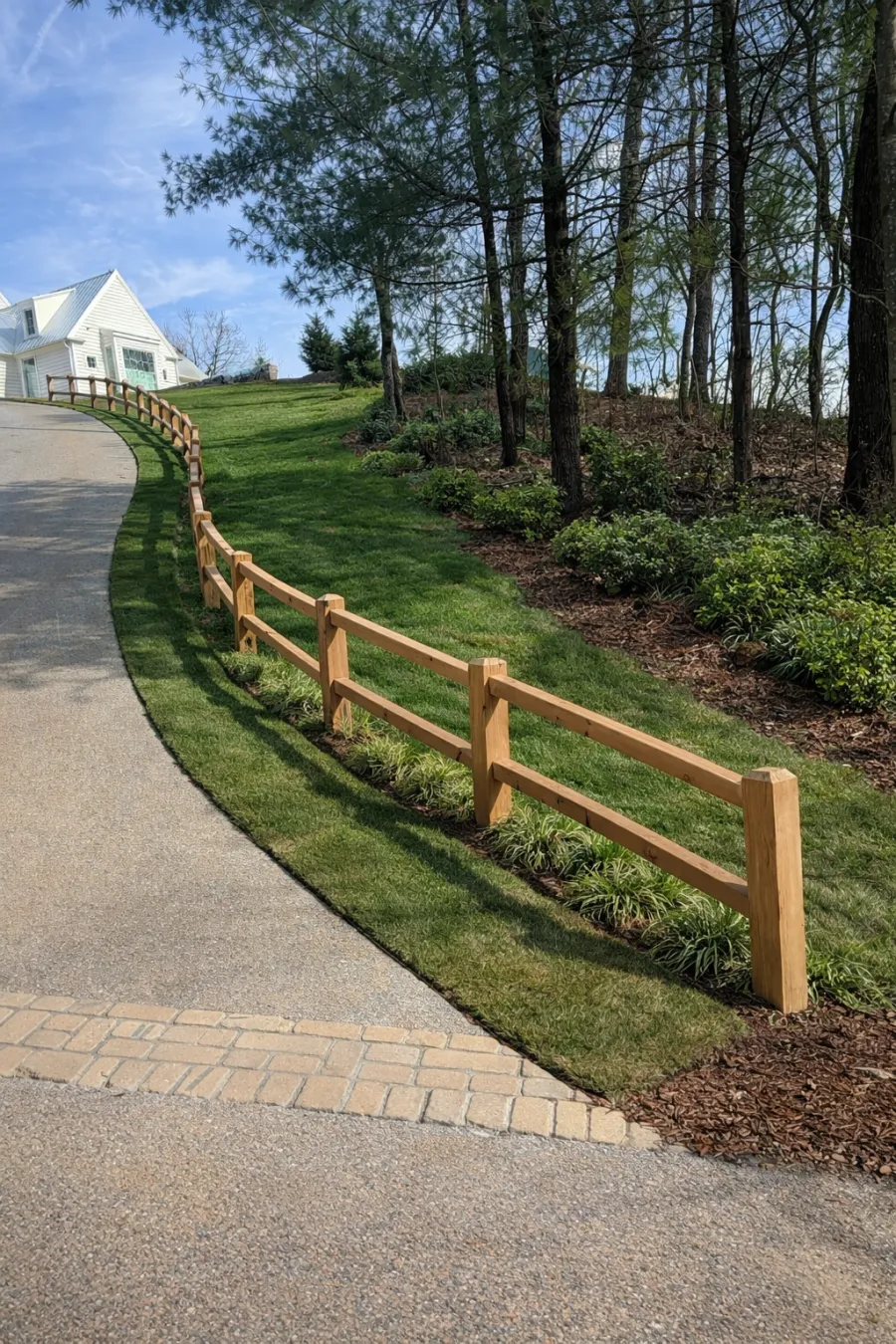 Curved concrete driveway bordered by a wooden fence and green lawn leading to a white house under blue sky.