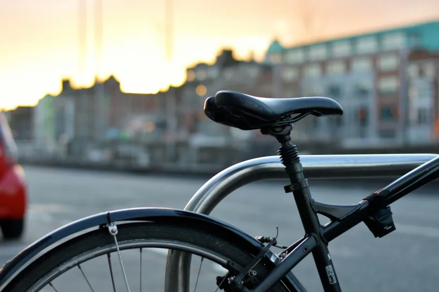 Close-up of a black bicycle seat and rear wheel locked to a metal rack with blurred urban buildings in the background at sunset