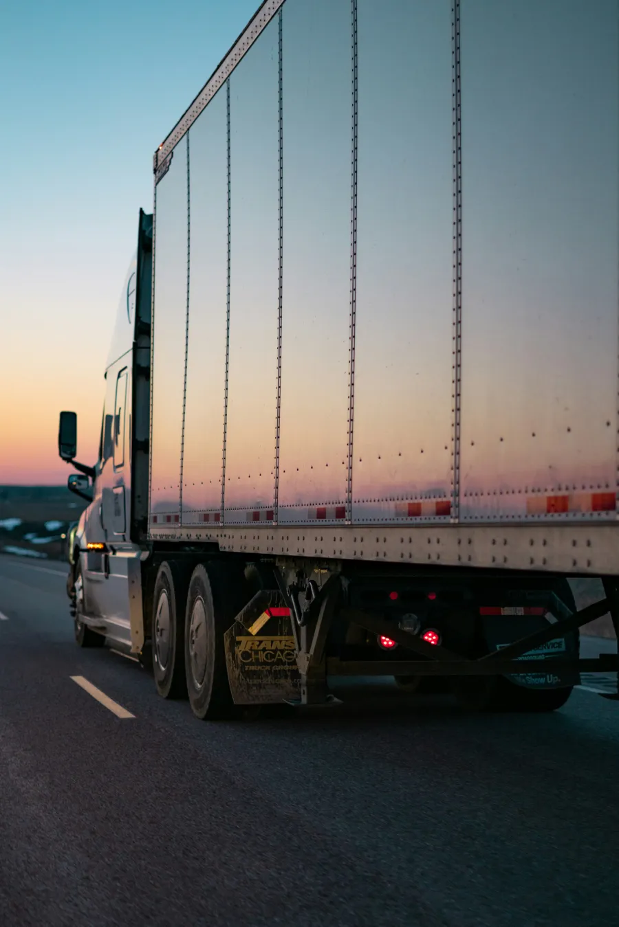Semi-truck driving on a highway at sunset with the trailer reflecting the colorful sky hues.