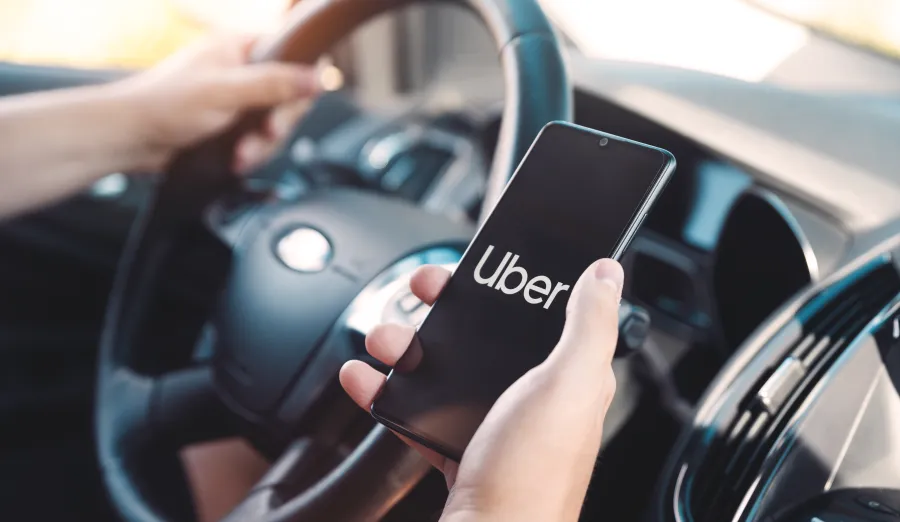 Person holding a smartphone with Uber app open inside a car, steering wheel visible in the background.