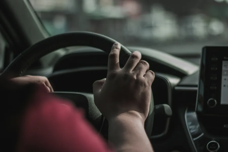 Close-up of a person's hand gripping a car steering wheel inside the vehicle, with dashboard in the background.