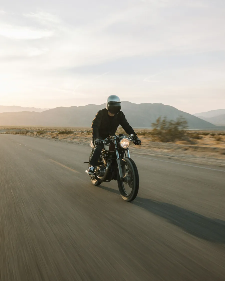 Motorcyclist wearing black gear rides on open road through desert landscape with mountains in distance