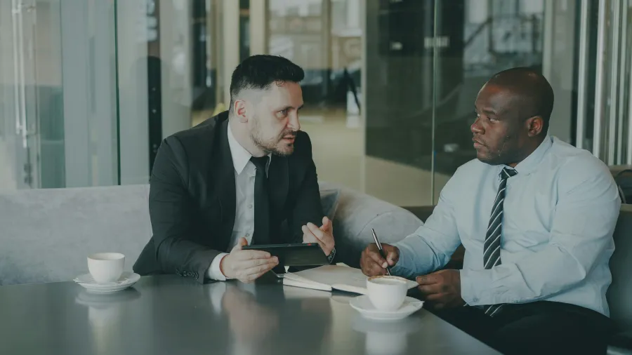 Two businessmen in formal attire having a serious discussion over coffee in a modern office lounge.