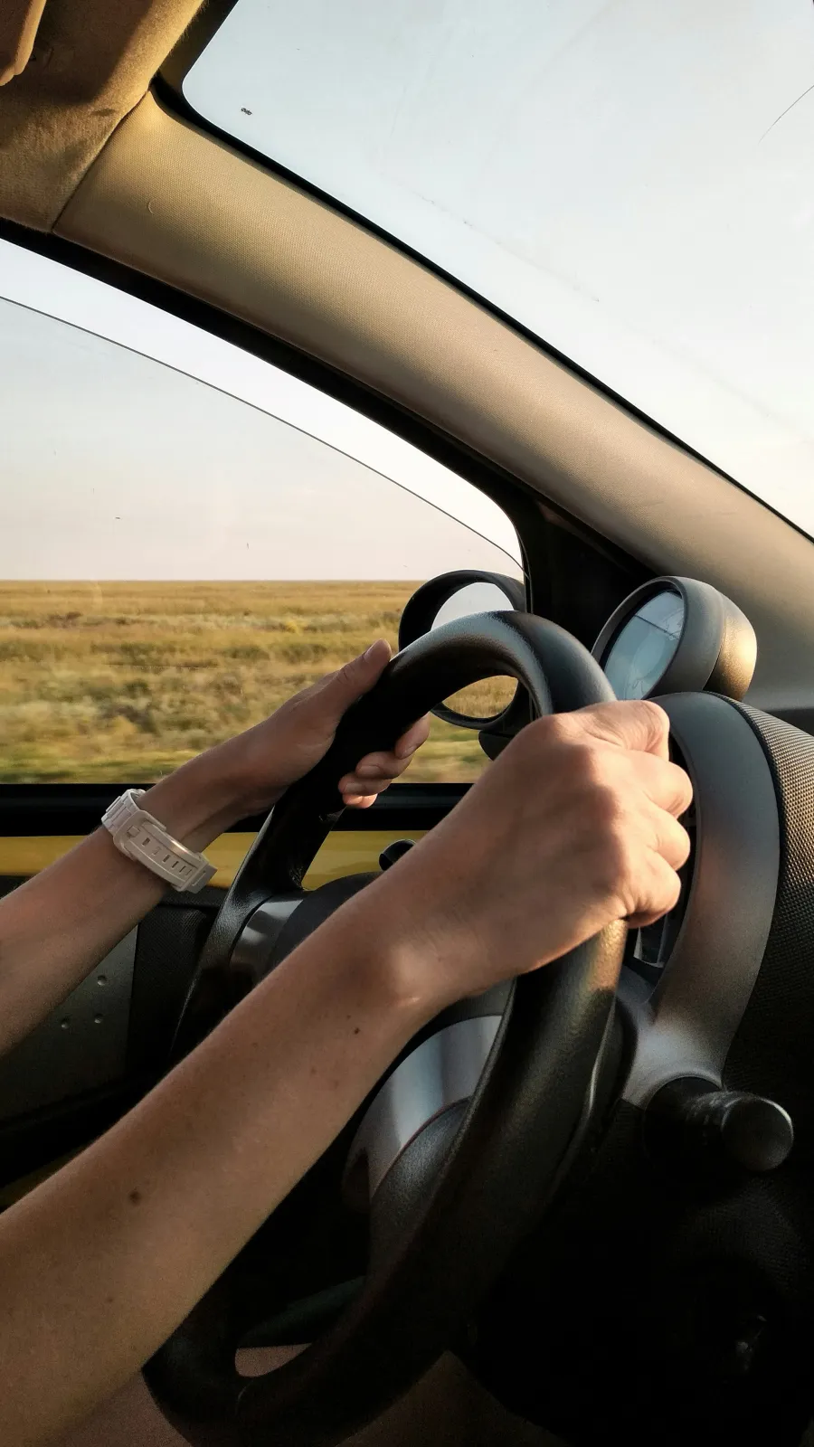 Person driving a car on an open road with hands on the steering wheel and a smartwatch visible.