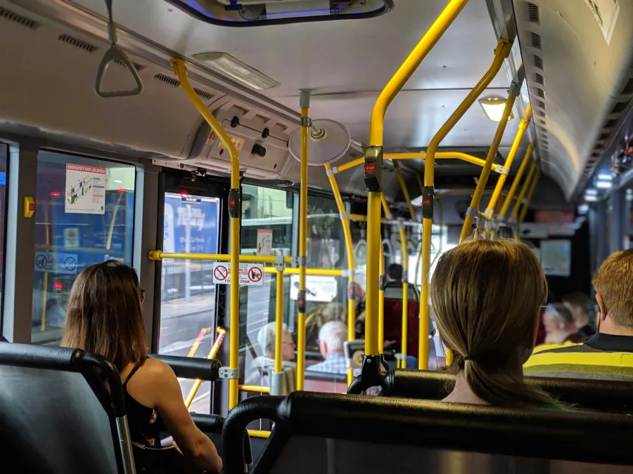 Passengers sitting inside a city bus with yellow poles and large windows during daytime.
