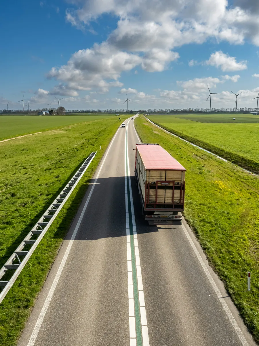 Truck driving on a rural highway surrounded by green fields and wind turbines under a partly cloudy sky.