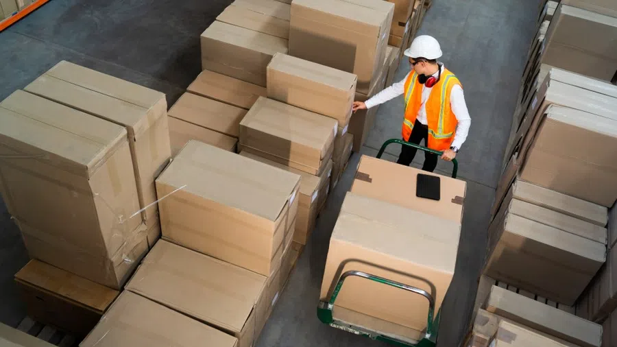 Warehouse worker in safety gear handling large cardboard boxes on a dolly in a storage facility.
