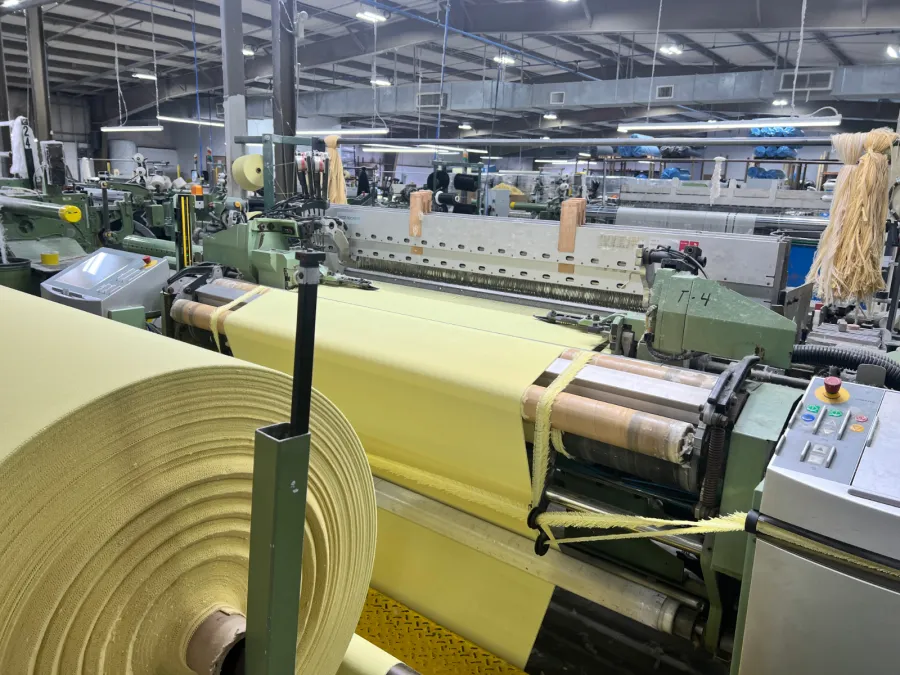 Industrial textile weaving machine producing wide yellow fabric rolls in a spacious factory setting.