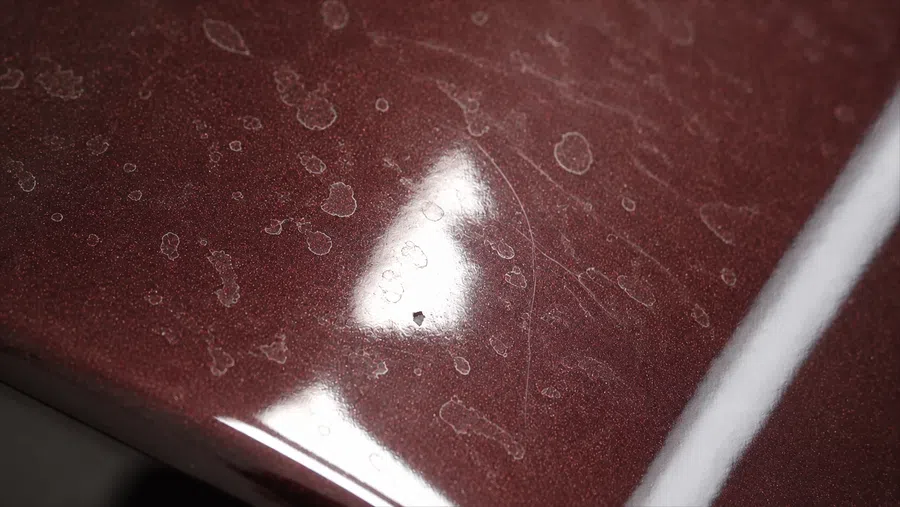 Close-up of dark red car paint with water spots, scratches, and light reflections on the surface.