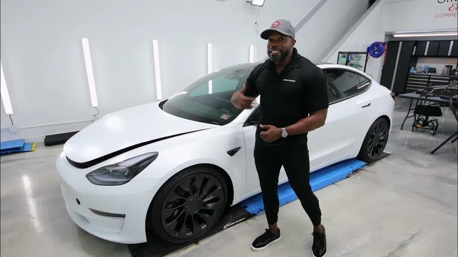 Man in black clothing and gray cap stands next to white Tesla Model 3 inside modern garage.