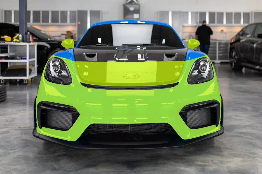 Front view of a green and blue sports car with black hood in a garage with tools and other cars.