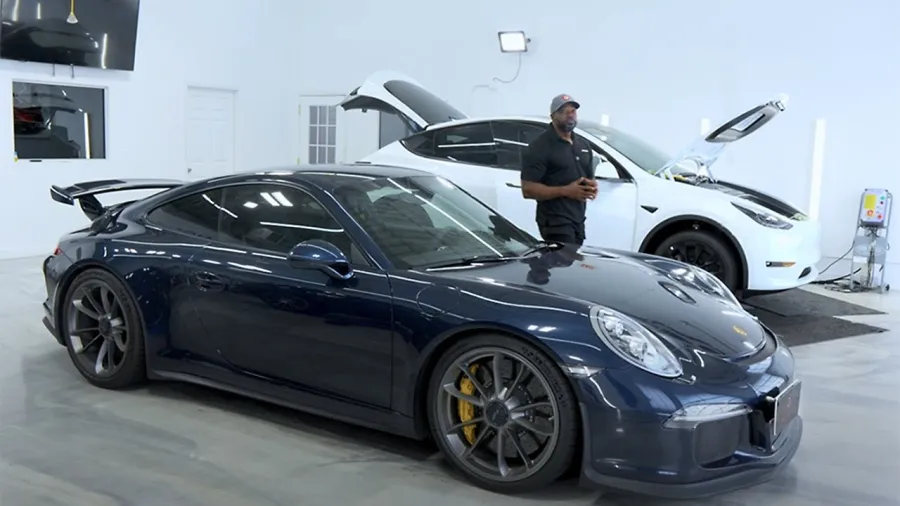 Black Porsche sports car and white Tesla SUV in garage with man standing beside Tesla with hood open.