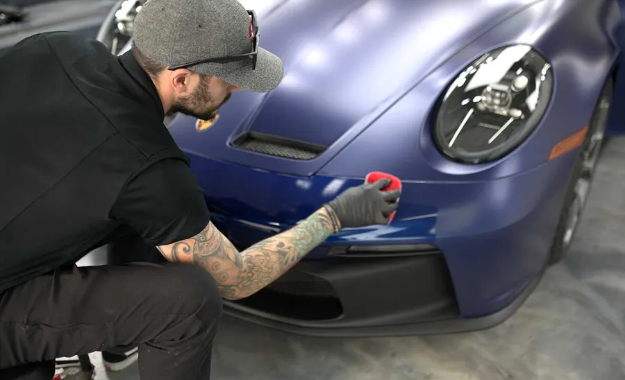 Man wearing a gray cap and gloves detailing the front bumper of a blue sports car in a garage.