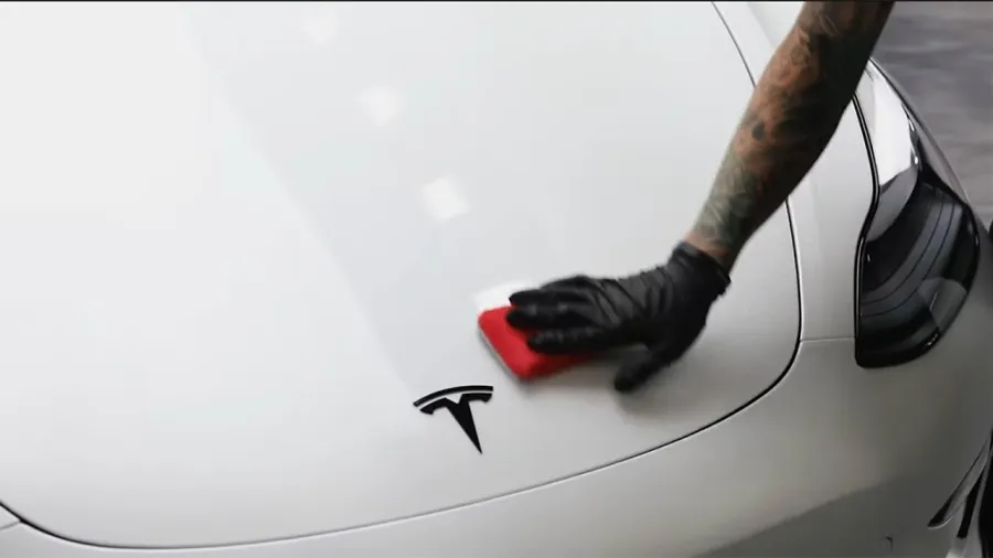 Person with tattooed arm and black glove polishing white Tesla car hood with a red cloth.