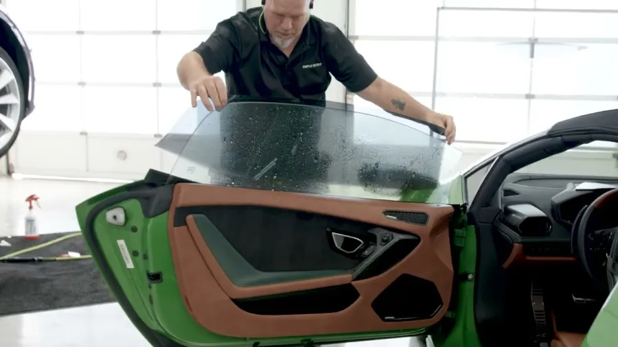 Technician installing tinted window film on the door of a green sports car inside a garage.
