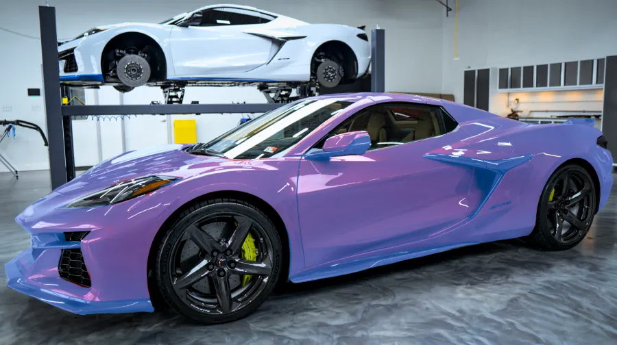 Purple sports car parked in a garage with a white car lifted on a hydraulic lift in the background.