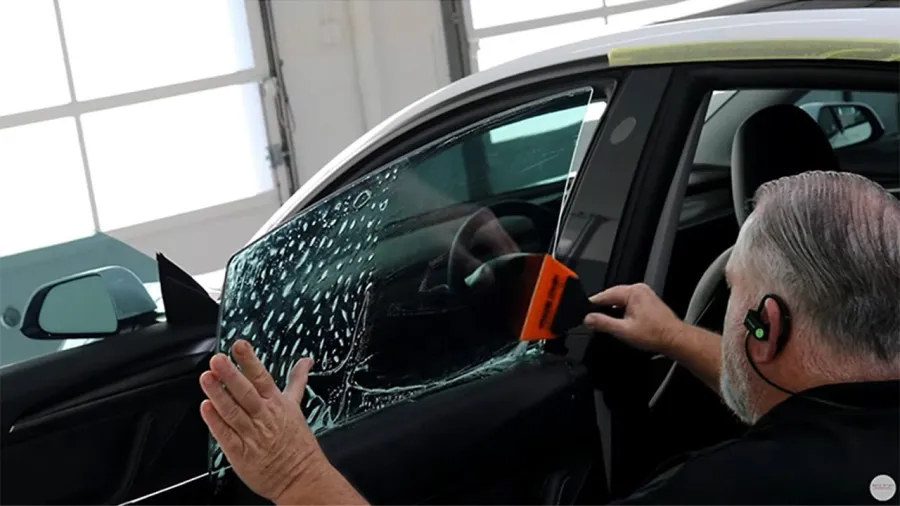 Man installing or tinting the side car window with tool and film inside a garage or workshop.
