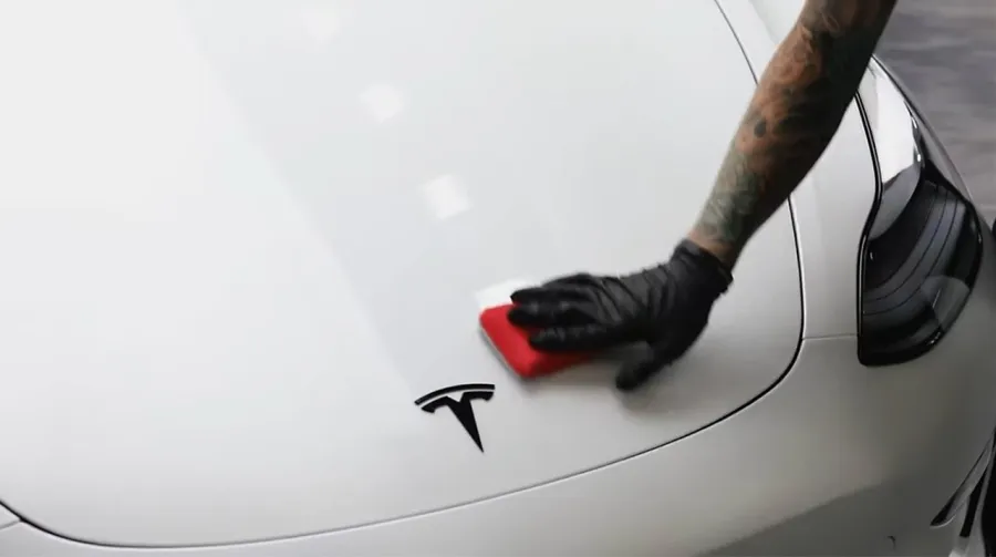 Person wearing black glove polishing white Tesla car hood with a red cloth near the Tesla logo.