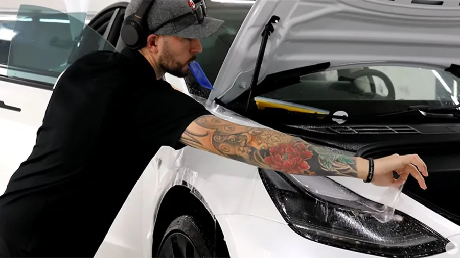 Man with tattooed arm applying protective film on white car's headlight inside a garage.