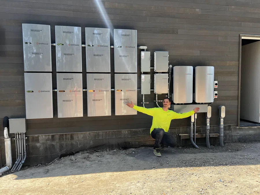 Man in neon yellow shirt kneeling by multiple Enphase electrical panels mounted on a brown exterior wall outdoors.