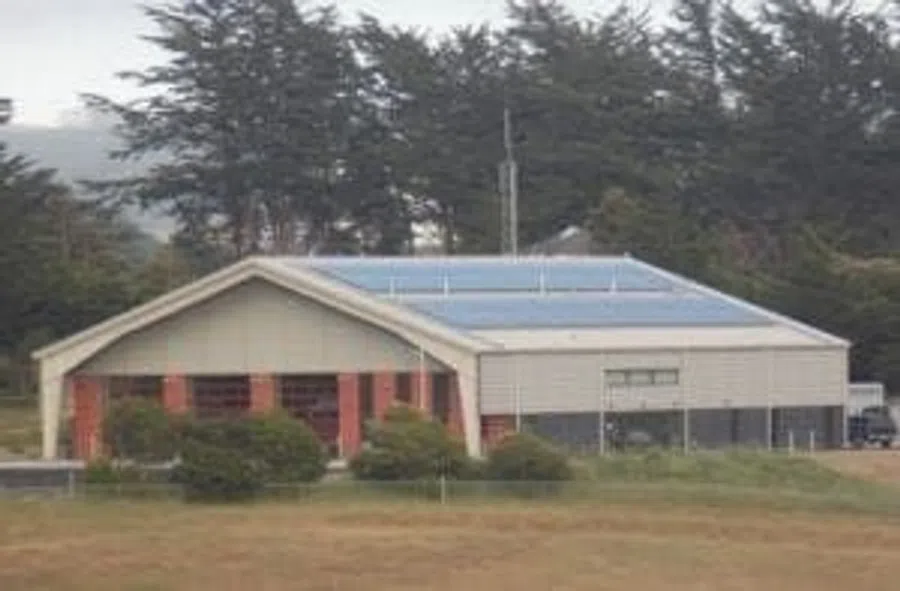 Large industrial building with solar panels on the roof surrounded by trees and grassy field.