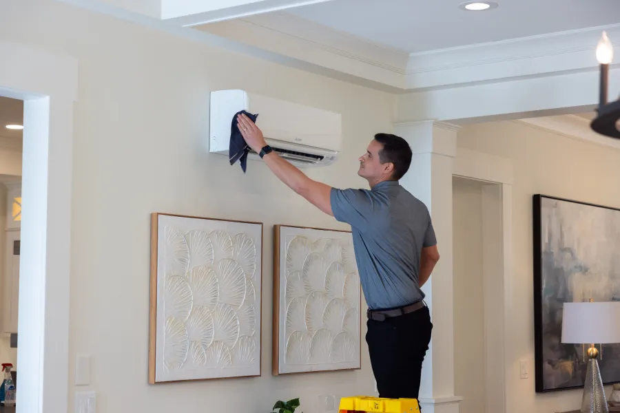 Man cleaning wall-mounted air conditioner unit with cloth in a modern living room with artwork and lamp