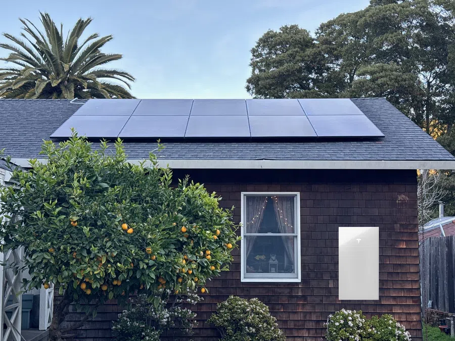 Solar panels on a house roof with a Tesla Powerwall battery mounted on the exterior wall beside a window and orange tree.
