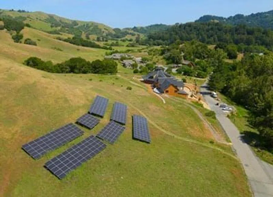 Solar panels installed on a grassy hillside near a rural building complex under clear sky.