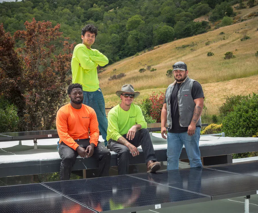 Four men in work attire posing near solar panels outdoors with hills and greenery in the background