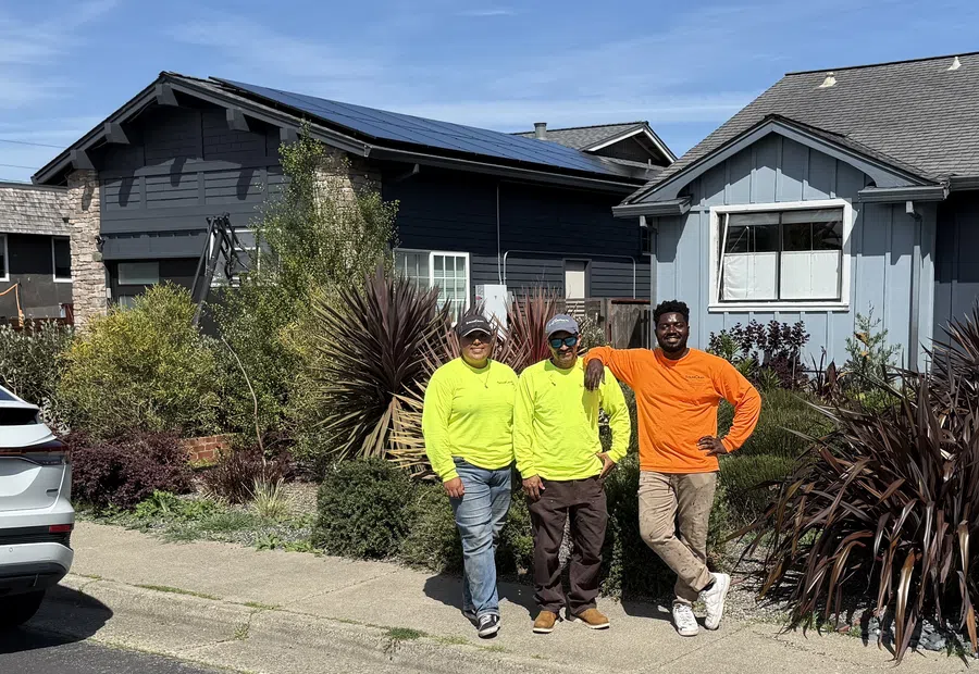 Three workers in bright shirts posing in front of a house with solar panels under clear blue sky