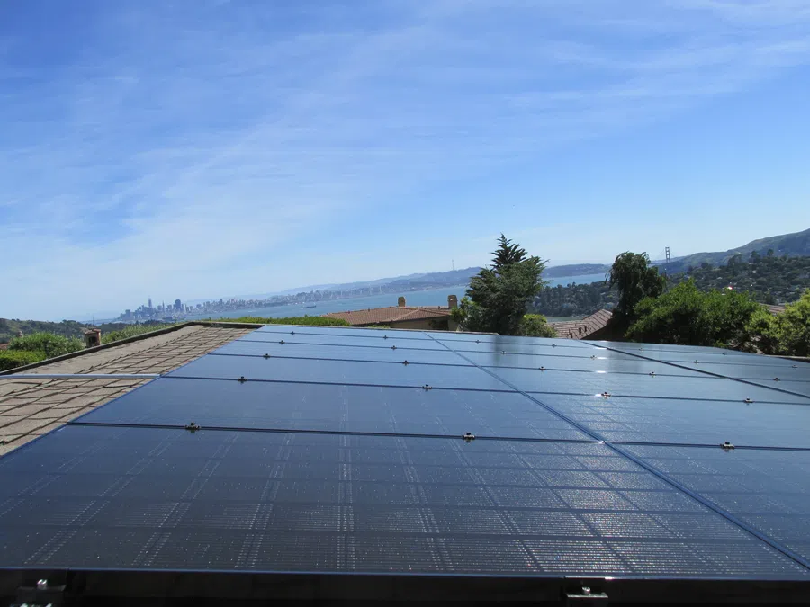 Solar panels installed on a rooftop with San Francisco skyline and Golden Gate Bridge in the background under blue sky.