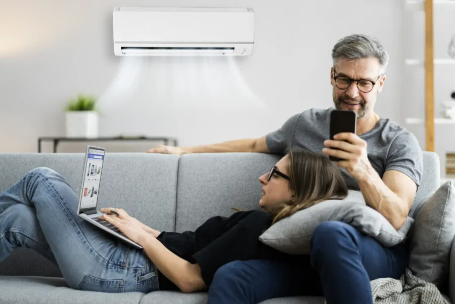 Couple relaxing on sofa using laptop and smartphone beneath a wall-mounted air conditioner blowing cool air.