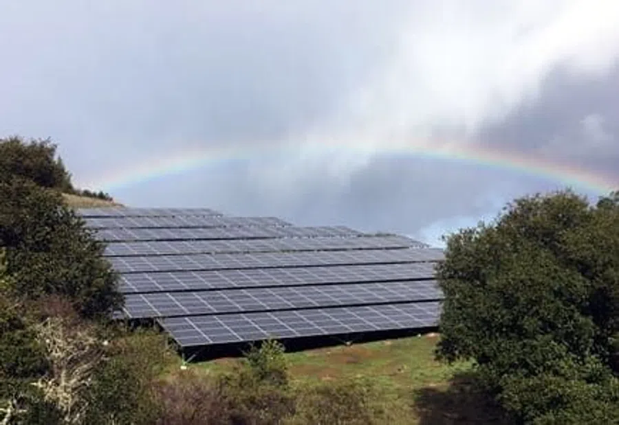 Solar panels on a green hillside with a rainbow and cloudy sky in the background.