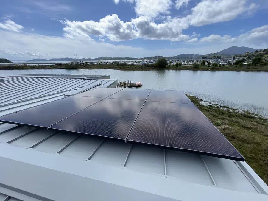 Solar panels mounted on metal roof overlooking water, boats, and distant hills under a blue sky with clouds