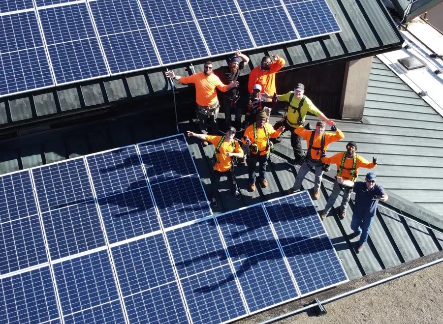 Group of workers in safety gear standing on roof with installed solar panels, waving at the camera.