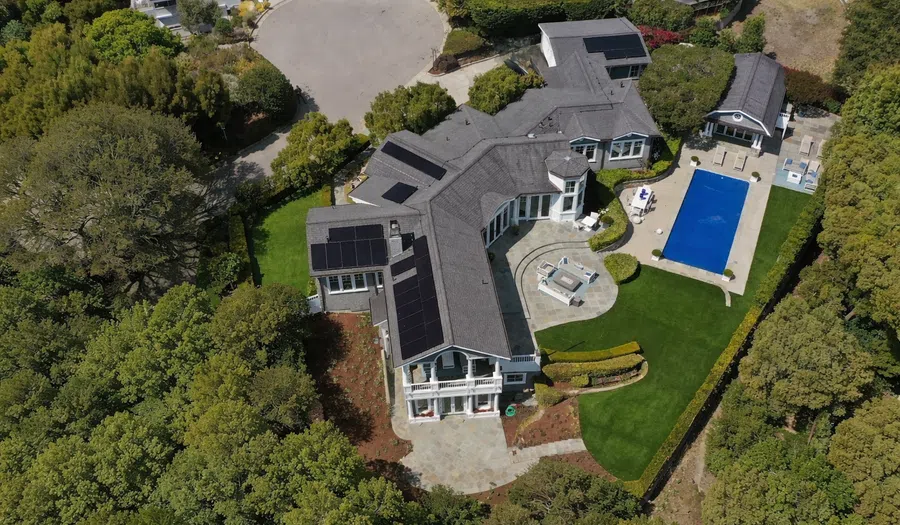 Aerial view of a modern large house with solar panels, swimming pool, patios, and manicured green lawn surrounded by trees.