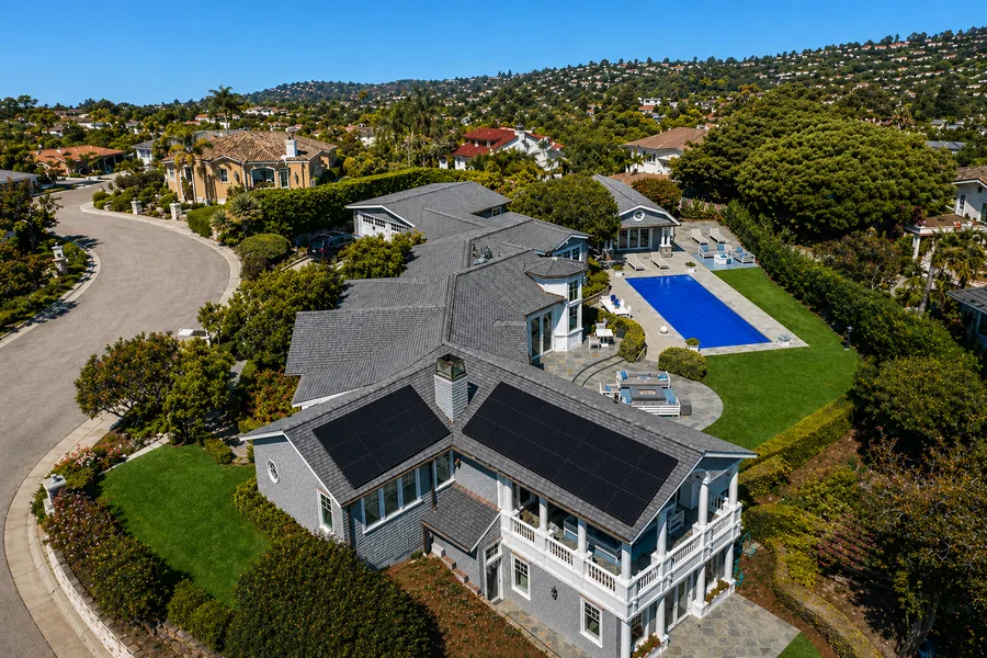 Aerial view of a large suburban house with solar panels on the roof and a backyard pool surrounded by greenery.