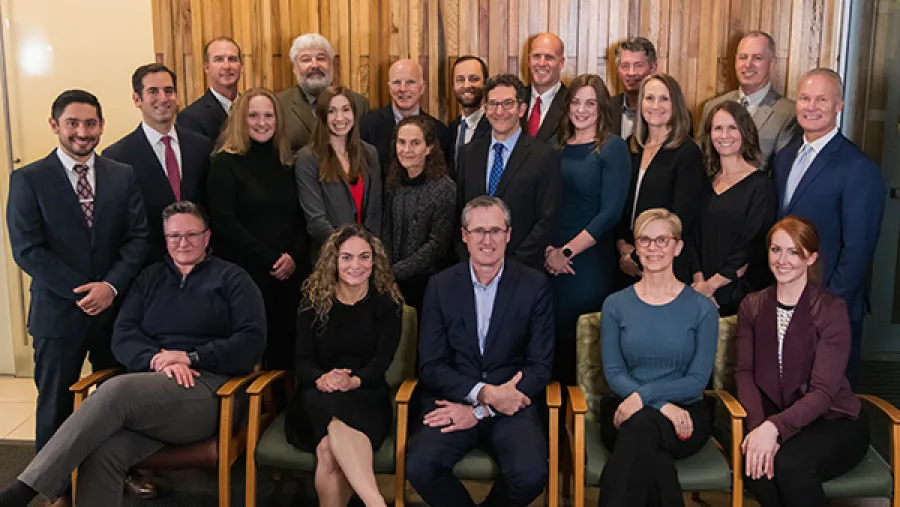 Group photo of 21 professional men and women posing indoors against wooden paneling, smiling at the camera.