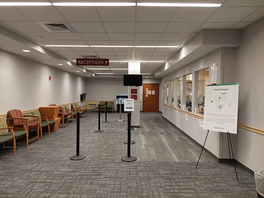 Empty medical facility lobby with seating, reception sign, and queue barriers under fluorescent lights.