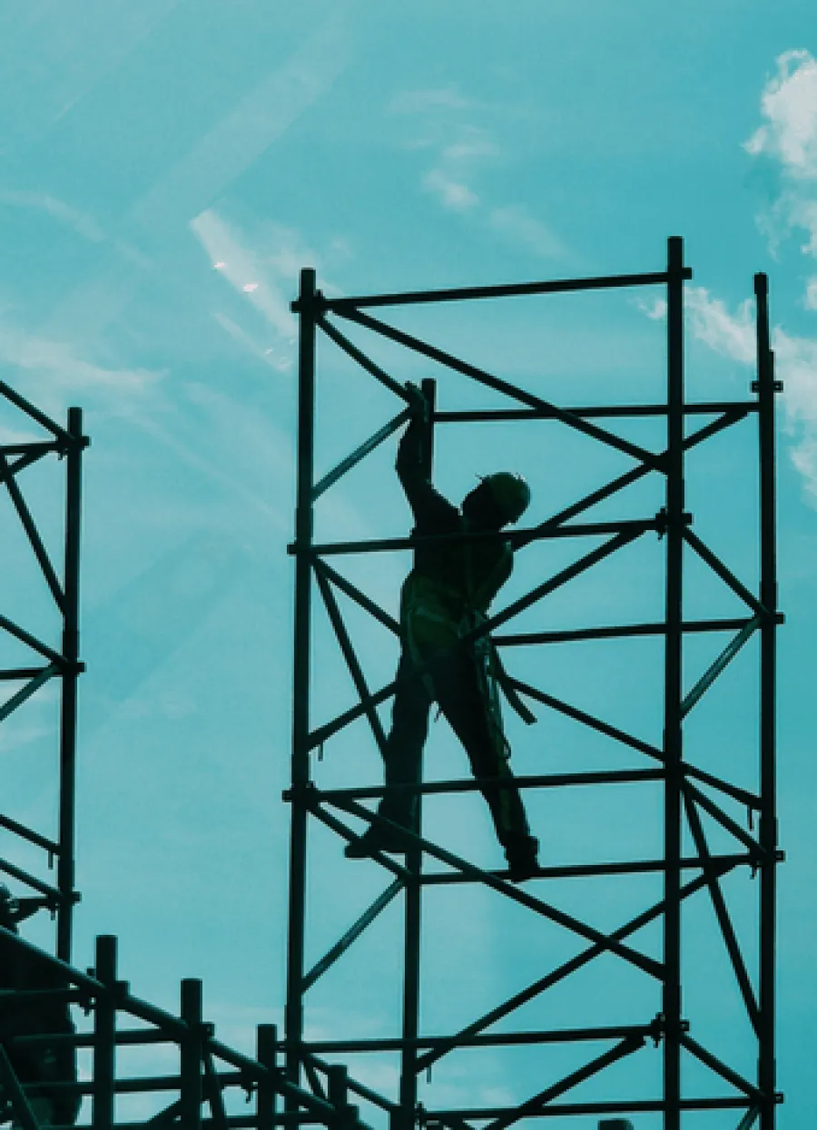 Silhouetted construction worker climbing scaffolding against a bright blue sky with scattered clouds.