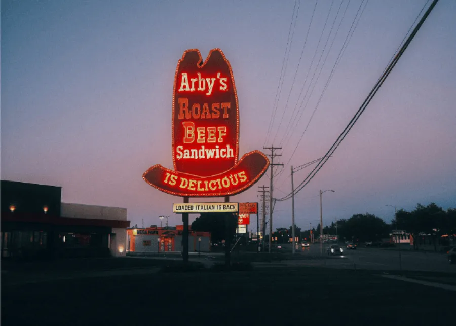 Arby's neon sign glowing at dusk advertising roast beef sandwiches and Loaded Italian sandwich.