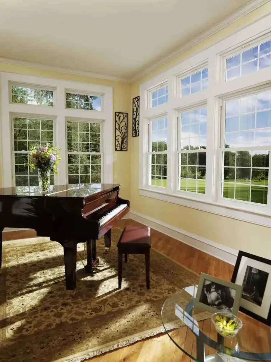 Bright music room with grand piano, large windows, floral rug, and glass table with framed photos and flowers.