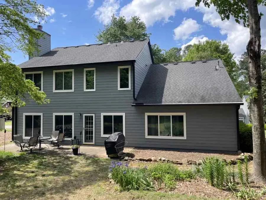 Two-story gray house with white trim, patio furniture, and garden under blue sky with scattered clouds