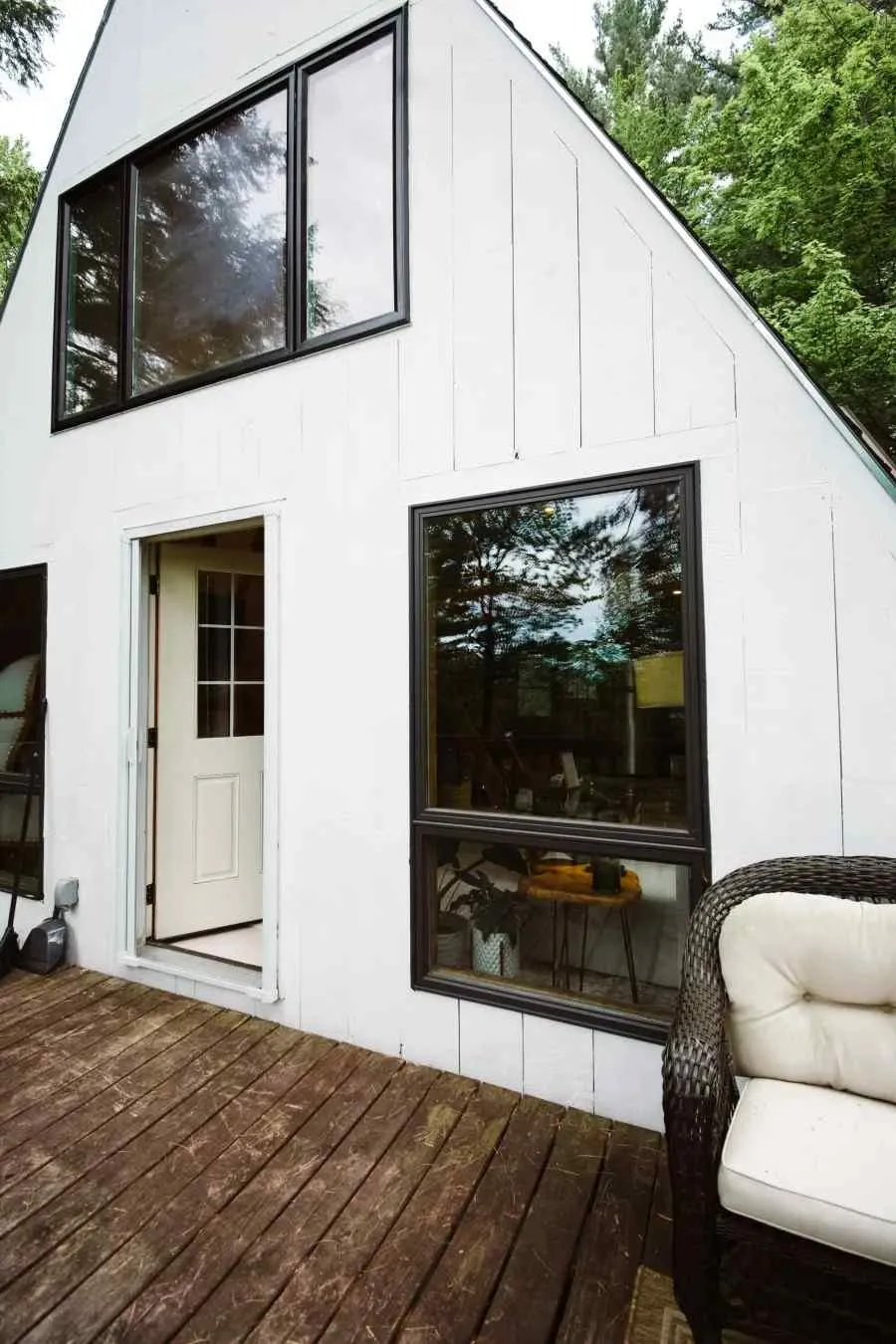 Cozy white cabin with large windows and an open door on a wooden deck surrounded by trees and nature.