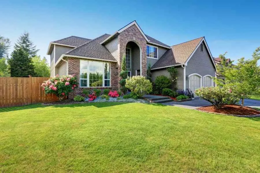 Two-story suburban house with brick and siding exterior, lush green lawn, and colorful flower beds under a clear blue sky