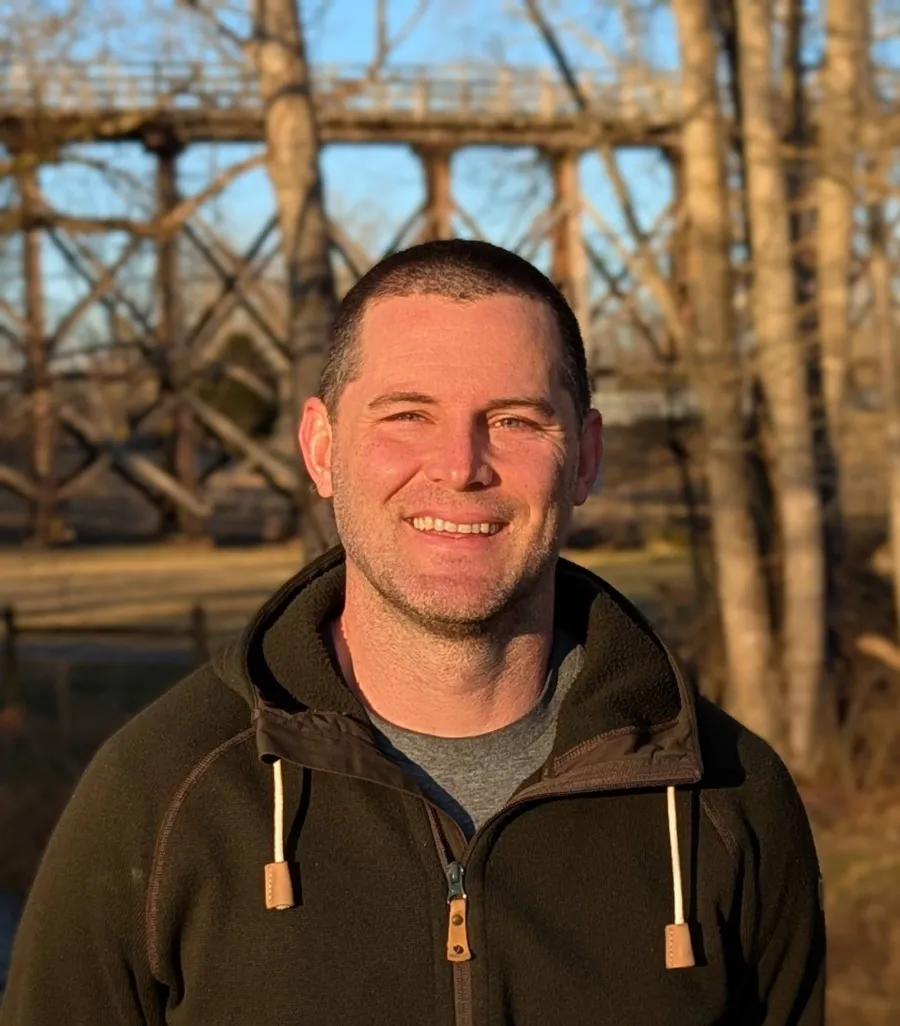 Smiling man in casual outdoor clothing stands in front of trees and wooden bridge on a sunny day.