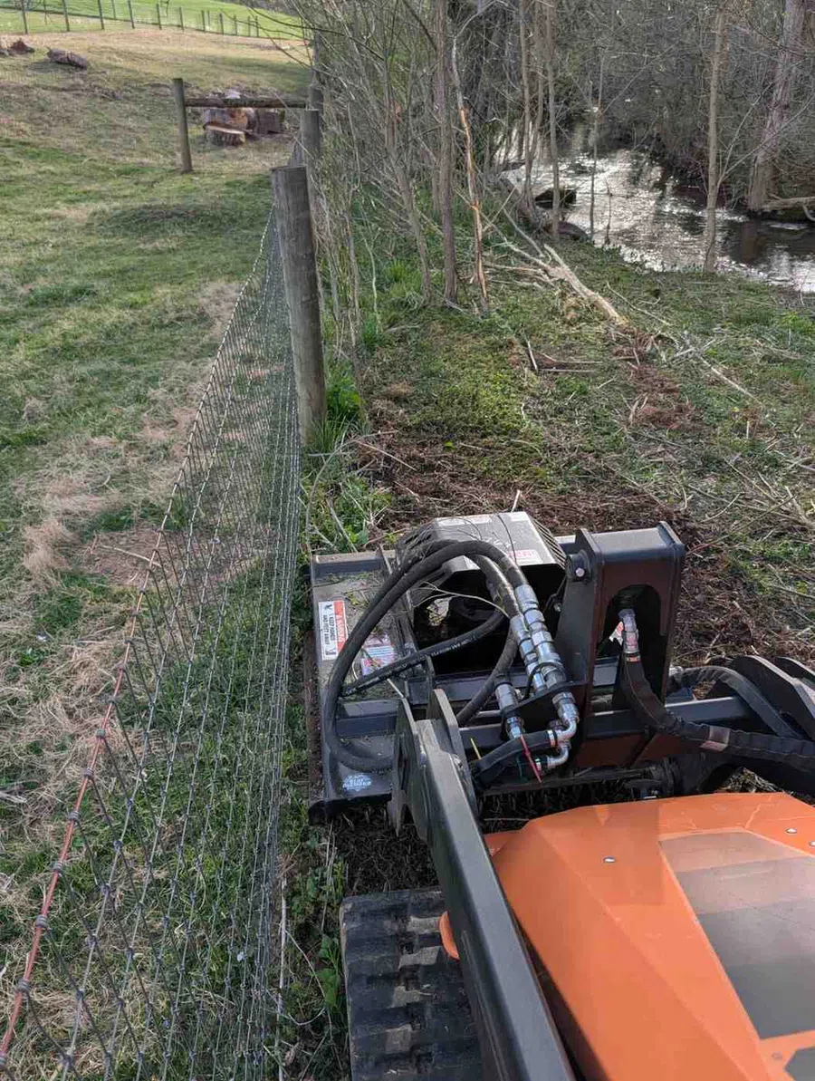 Orange excavator clearing brush along wire fence beside a creek in a rural field with trees and grass.