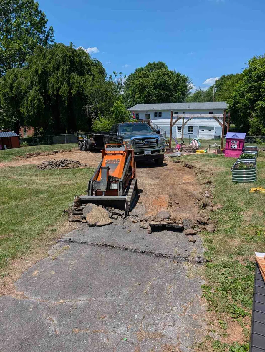 Mini excavator breaking asphalt driveway with truck and playground in backyard during sunny day