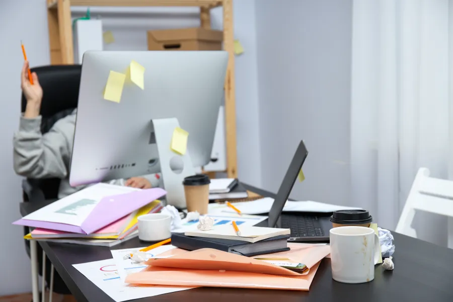 Busy workspace with cluttered desk, multiple laptops, coffee cups, notebooks, and sticky notes in an office setting