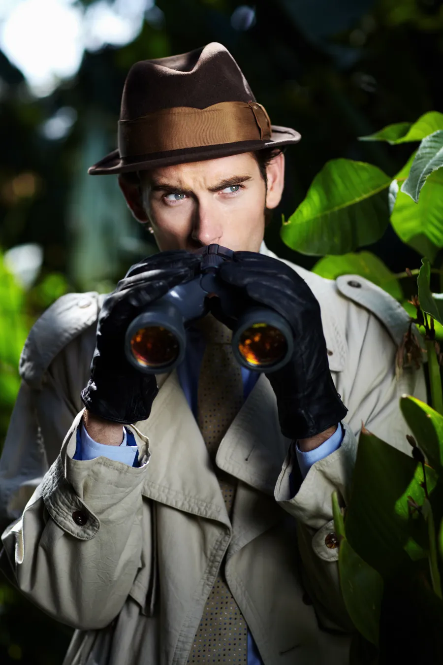 Man in trench coat and hat using binoculars while hiding among green foliage in a forest or jungle.